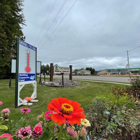 A photo showing a sign for the Lakes Country Library next to a fundraising sign with pink, yellow, and orange flowers.